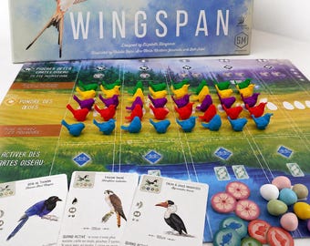 Puede incluir: Un juego de mesa Wingspan que incluye una caja grande con la ilustración de un ave, un tablero con filas de figuras de aves de plástico en verde, amarillo, morado, rojo y azul, tres cartas de aves y fichas redondas de huevos en tonos pastel.