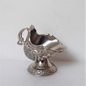 May include: A silver gravy boat with a handle and a pedestal base. The boat has a detailed grape and leaf design on the side. The handle has a decorative swirl design. The base has a floral pattern.