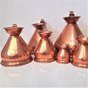 May include: Set of six copper measuring jugs with handles. The jugs are labelled with different measurements: pint, 1/2 pint, quart, half gallon, and 1 gallon.