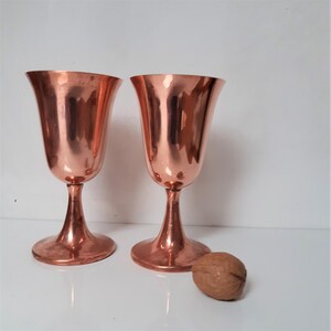 May include: Two copper goblets with a flared bowl and a stemmed base. The goblets are polished and have a warm, metallic sheen.