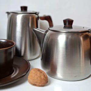 May include: Two stainless steel teapots with dark brown wooden handles and lid knobs, accompanied by a dark brown ceramic cup and saucer set and a single whole walnut. The teapots feature a polished metallic finish and a classic rounded shape.