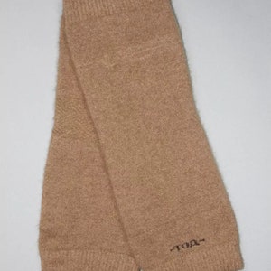 May include: A pair of tan-colored leg warmers made of a soft, knitted material. The leg warmers are folded and feature a ribbed cuff at the top and bottom. The text "-TDA-" is embroidered near the bottom of one leg warmer.