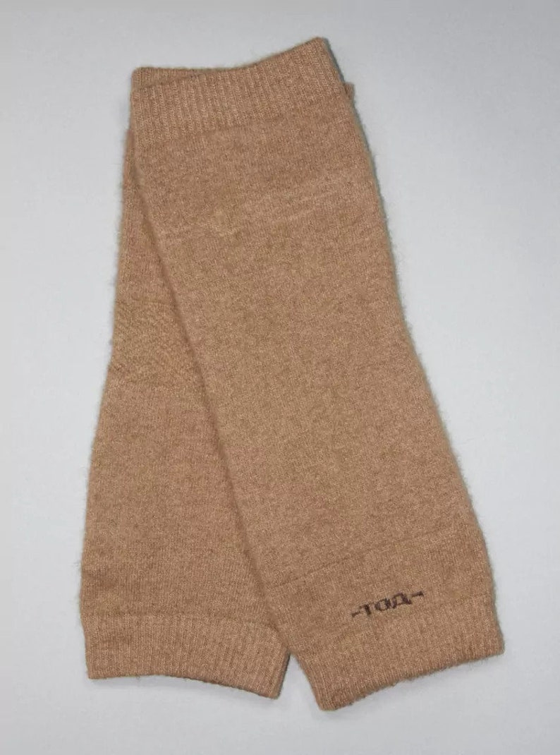 May include: A pair of tan-colored leg warmers made of a soft, knitted material. The leg warmers are folded and feature a ribbed cuff at the top and bottom. The text "-TDA-" is embroidered near the bottom of one leg warmer.