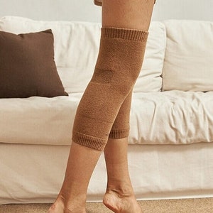 May include: A pair of brown knee warmers made of a soft knit material. The warmers are shown on a person's legs, with ribbed cuffs at the top and bottom. The background includes a white sofa and a brown pillow.