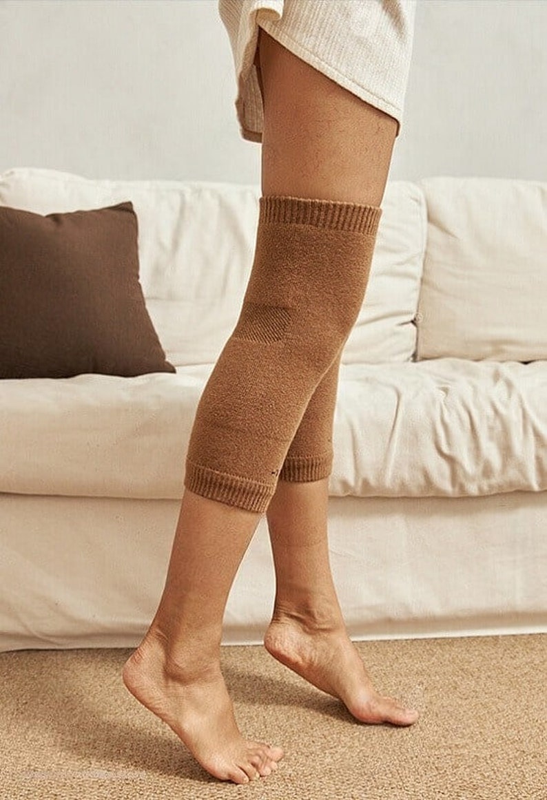 May include: A pair of brown knee warmers made of a soft knit material. The warmers are shown on a person's legs, with ribbed cuffs at the top and bottom. The background includes a white sofa and a brown pillow.