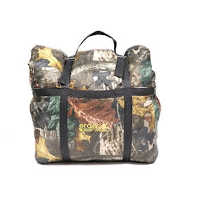 Può includere: Un sacco a pelo con motivo mimetico, cinturini neri e una maniglia. Il sacco è piegato e compresso, e il nome del marchio "ordonez SHEEP WOOL" è visibile sul davanti.