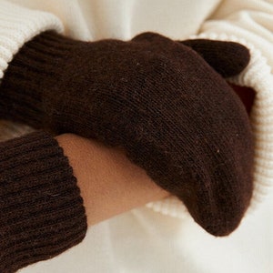 May include: Close-up of dark brown gloves made of a soft, knitted material. The gloves are worn over a cream-colored sweater, with the cuff of the sweater visible. The gloves appear warm and comfortable, suitable for cold weather.