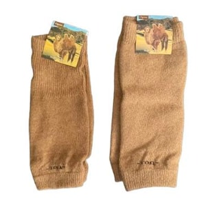 May include: Two tan-colored leg warmers with a ribbed cuff and a small tag featuring a camel image. The leg warmers are made of a soft, knitted material. The leg warmers are displayed on a white background.
