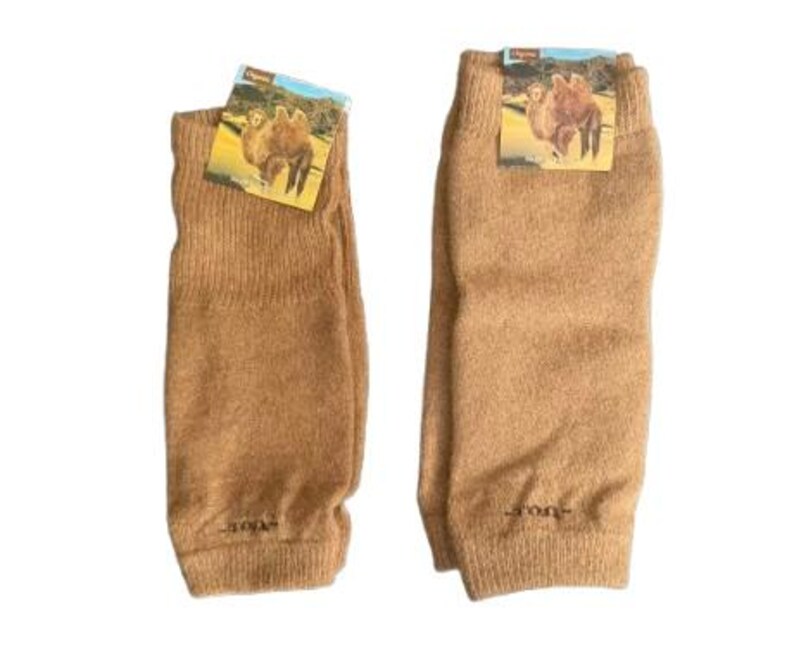 May include: Two tan-colored leg warmers with a ribbed cuff and a small tag featuring a camel image. The leg warmers are made of a soft, knitted material. The leg warmers are displayed on a white background.