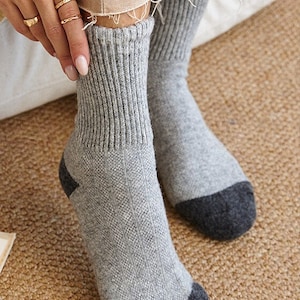 May include: A pair of gray socks with dark gray toes and heels. The socks have a ribbed cuff and a textured pattern on the front. The person is wearing gold rings. The socks are on a brown woven surface.