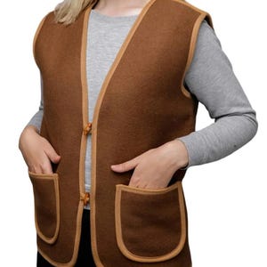 Puede incluir: Un chaleco marrón con dos bolsillos delanteros y un ribete marrón claro. El chaleco tiene cierre de botones y está hecho de un material suave y texturizado. La modelo lleva una camiseta gris de manga larga.