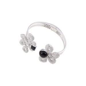 Puede incluir: Un brazalete de plata con dos acentos de flores de piedras preciosas negras. Las flores están hechas de piedras de circonita cúbica transparentes.