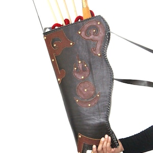 May include: A black leather quiver with brown leather accents and gold studs. The quiver is decorated with intricate designs and has a strap for carrying. It is holding a set of arrows and a bow.