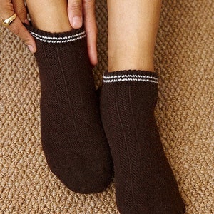 Puede incluir: Un par de calcetines tobilleros marrones con un diseño acanalado. Los calcetines tienen una banda a rayas blancas y negras en la parte superior. Los calcetines están siendo sostenidos por una persona.