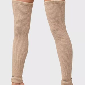 May include: Beige ribbed knit leg warmers. These knee-high leg warmers are made of a soft, textured material. The leg warmers have a rolled cuff at the top and bottom. The bottom cuff has a small black logo. These leg warmers are a neutral color.