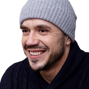 May include: A light gray ribbed knit beanie. The beanie is worn by a person smiling, with a dark blue sweater. The background is white.