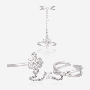 Puede incluir: Un anillo de plata con un diseño de nudo y una cadena que conecta con un segundo anillo. La cadena tiene una pequeña piedra transparente.