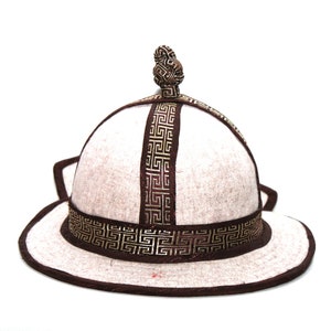 Può includere: Cappello tradizionale con corona arrotondata e tesa larga. Il cappello è principalmente bianco sporco con finiture marroni e motivi geometrici dorati decorativi. Un nodo decorativo si trova sulla sommità della corona. Il cappello ha paraorecchie.