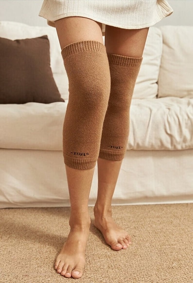 May include: Knee-high brown leg warmers with ribbed cuffs. The warmers are made of a soft, knitted material and feature a small embroidered detail near the cuff. The person is standing barefoot on a beige carpet.