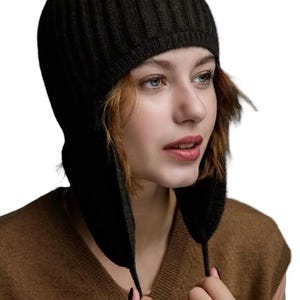 Può includere: Un cappello nero a maglia con paraorecchie. Il cappello ha una trama a coste e copre la testa e le orecchie, con lacci pendenti. La persona indossa un maglione marrone.
