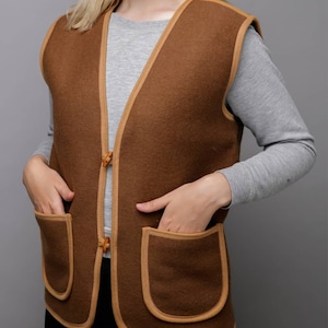 May include: A brown sleeveless vest with a tan trim and two large pockets. The vest has a button closure.