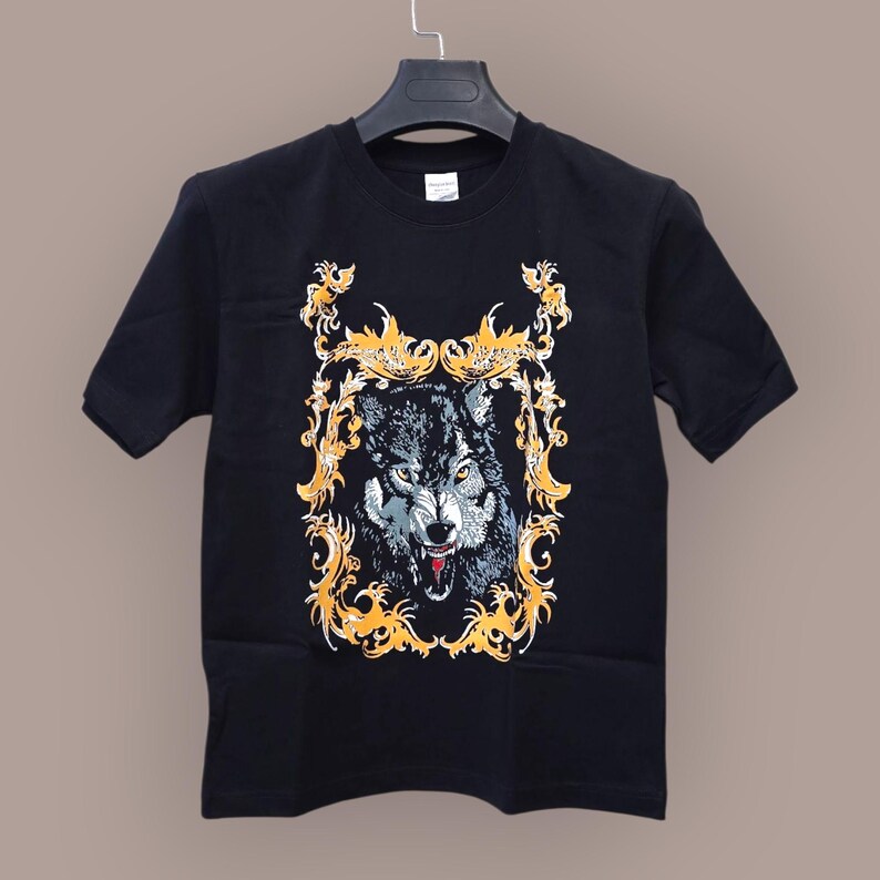 May include: Black t-shirt featuring a detailed illustration of a wolf with an open mouth, set within a decorative gold frame. The wolf's fur is rendered in shades of gray and white, with red accents on the tongue. The shirt is hanging on a black hanger.