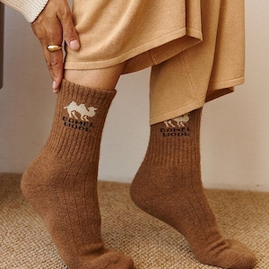 May include: A pair of brown ribbed socks with the text "CAMEL WOOL" and a camel graphic. The socks are worn with tan pants and a cream-colored sweater. The socks are made of a soft, warm material.