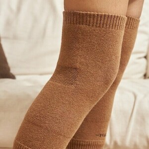 May include: Close-up of two brown knee sleeves. The sleeves are made of a knitted material and cover the knees and upper shins. The sleeves have a ribbed cuff at the top and bottom. The background is a white sofa.