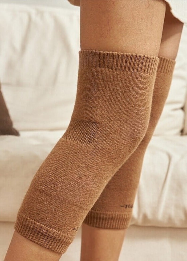 May include: Close-up of two brown knee sleeves. The sleeves are made of a knitted material and cover the knees and upper shins. The sleeves have a ribbed cuff at the top and bottom. The background is a white sofa.