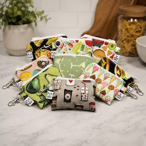 May include: A collection of small rectangular fabric pouches with silver-toned metal lobster claw clasps. Each pouch features a unique colorful print including patterns of tacos, donuts, avocados, ice cream cones, coffee drinks, and martini glasses.