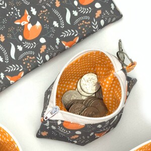 Puede incluir: Una peque&ntilde;a bolsa de cremallera naranja y gris con estampado de zorro. La bolsa est&aacute; abierta y llena de monedas.