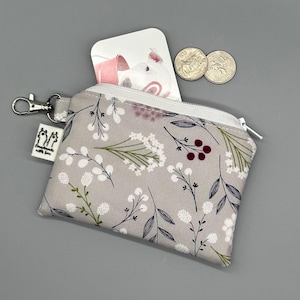 May include: A gray floral coin purse with a white zipper, a silver clasp, and a small tag that says "handmade with love." The purse contains a card with a pig illustration and two quarters.