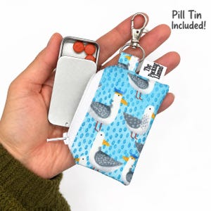 May include: A light blue zippered pouch featuring a repeating seagull pattern, attached to a silver metal swivel clasp. A rectangular metal pill tin containing orange pills is tucked inside. A white tag reads The Packing Llamas.