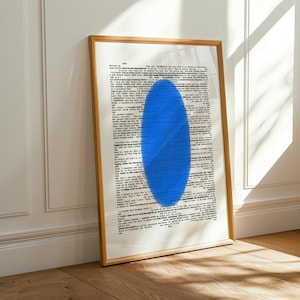 May include: A framed art print featuring a page of printed text with a large, vibrant blue oval shape in the center. The frame is a light brown color, and the artwork is leaning against a white wall and a wooden floor.