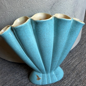 May include: A turquoise ceramic vase with a fan-shaped design. The vase has a speckled texture and a scalloped rim with a cream-colored interior. A gold bird is on the base. The vase is approximately 8 inches tall.