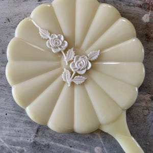 May include: A vintage hand mirror in a pale yellow hue. The mirror features a fan-shaped design with a scalloped edge and a handle. Two white floral embellishments adorn the face of the mirror, adding a touch of elegance.