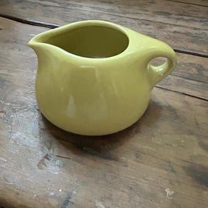 May include: A small, solid light yellow ceramic creamer pitcher with a smooth, glossy finish. The pitcher features a rounded body, a wide pouring spout on one side, and a single integrated handle on the opposite side for easy gripping and pouring.