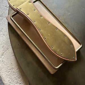 May include: A vintage shoe stretcher made of wood and brass. The stretcher has a curved shape with a brass plate on top and is secured with rivets. The handle is made of brass and is rectangular. The shoe stretcher is on a round brass table.