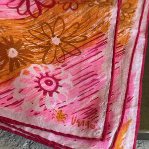 May include: A vintage floral scarf in shades of pink, orange, and brown. The scarf features a repeating pattern of stylized flowers and a handwritten signature in the corner. The edges are trimmed with a darker pink border.