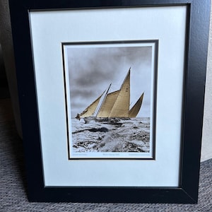 May include: A black and white photograph of a sailboat with sails billowing in the wind. The sailboat is in the middle of the ocean with waves crashing around it. The photo is framed in a black frame with a white mat.