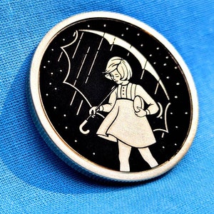 May include: A black and white circular pin with a girl holding an umbrella in the rain. The girl is wearing a dress and has her hair pulled back. The background is black with white raindrops and stars.