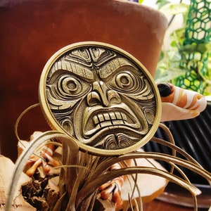 May include: A gold-colored metal coin with a detailed carving of an angry face. The face has a wide mouth, large eyes, and a furrowed brow. The coin is sitting on a plant with long, thin leaves.