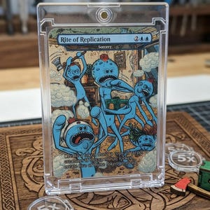 May include: A clear plastic card holder containing a Magic: The Gathering card. The card features a cartoon illustration of blue creatures. The card is labeled "Rite of Replication" and "Sorcery". The holder sits on a wooden surface with etched designs and acrylic tokens.