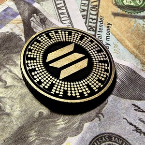 May include: A gold and black cryptocurrency coin with a stylized three-bar logo on a black background. The coin is on top of a US dollar bill.