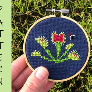May include: A close-up of a finished cross-stitch embroidery project depicting a Venus flytrap plant with a red flower and a black and white fly. The embroidery is framed in a round wooden hoop.