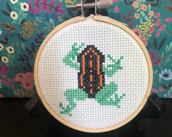 May include: A cross-stitch embroidery hoop with a green frog with an orange and black shell. The hoop is on a black stand.