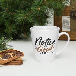 May include: A white ceramic latte mug featuring the black and brown printed text Notice THE Good STUFF with a small TGL logo. The mug has a tapered shape with a curved handle and a smooth finish, displaying the design clearly on the front surface.