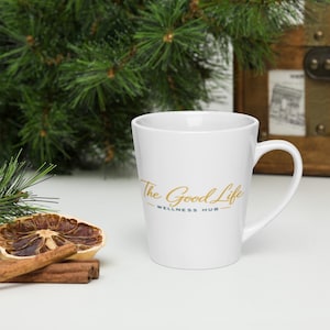 May include: A white ceramic latte mug featuring a gold script logo that reads The Good Life and dark blue text below that reads WELLNESS HUB. The mug has a tapered shape with a curved handle on the right side and a smooth, glossy finish.