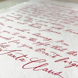 Può includere: Primo piano di un foglio di carta bianca con scritta a mano in inchiostro rosso. La scritta include le parole "Janine", "benedizione" e "Santa Claus". La carta ha una superficie strutturata e la scritta è in corsivo.