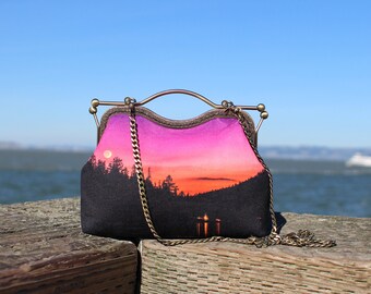 May include: A small, black fabric purse with a gold chain strap. The purse has a sunset scene printed on the front with a pink and orange sky and a silhouette of trees.